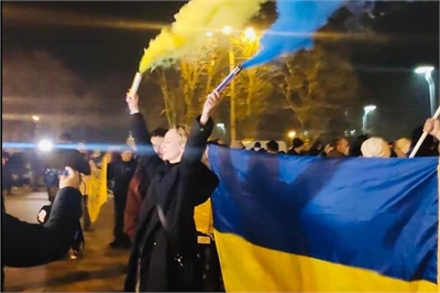 [ai] A group of people gathered at night, holding a large Ukrainian flag while some wave blue and yellow smoke flares. There are lights in the background and a crowd visible.